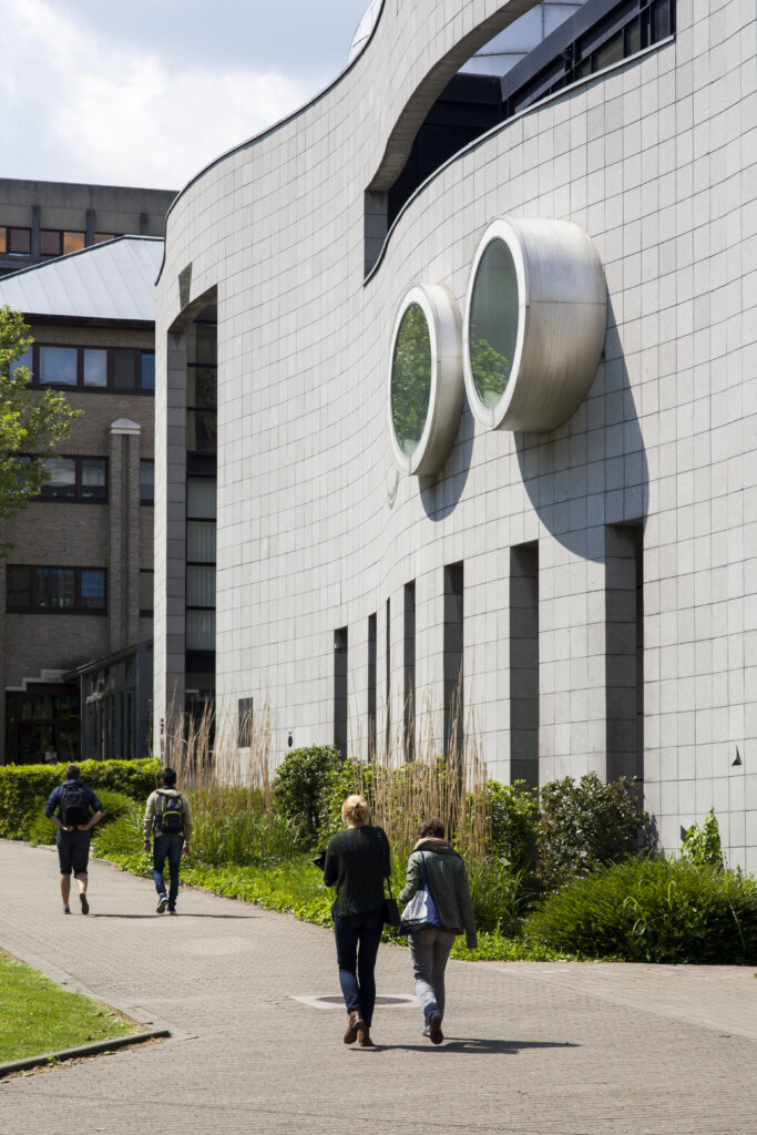 Vue extérieure du campus d'Erasme de l'Université libre de Bruxelles (ULB), mettant en avant une façade moderne avec des fenêtres circulaires emblématiques et une allée bordée de verdure. Des étudiants, sacs à dos en main, se dirigent vers le bâtiment, reflétant une atmosphère active et académique.